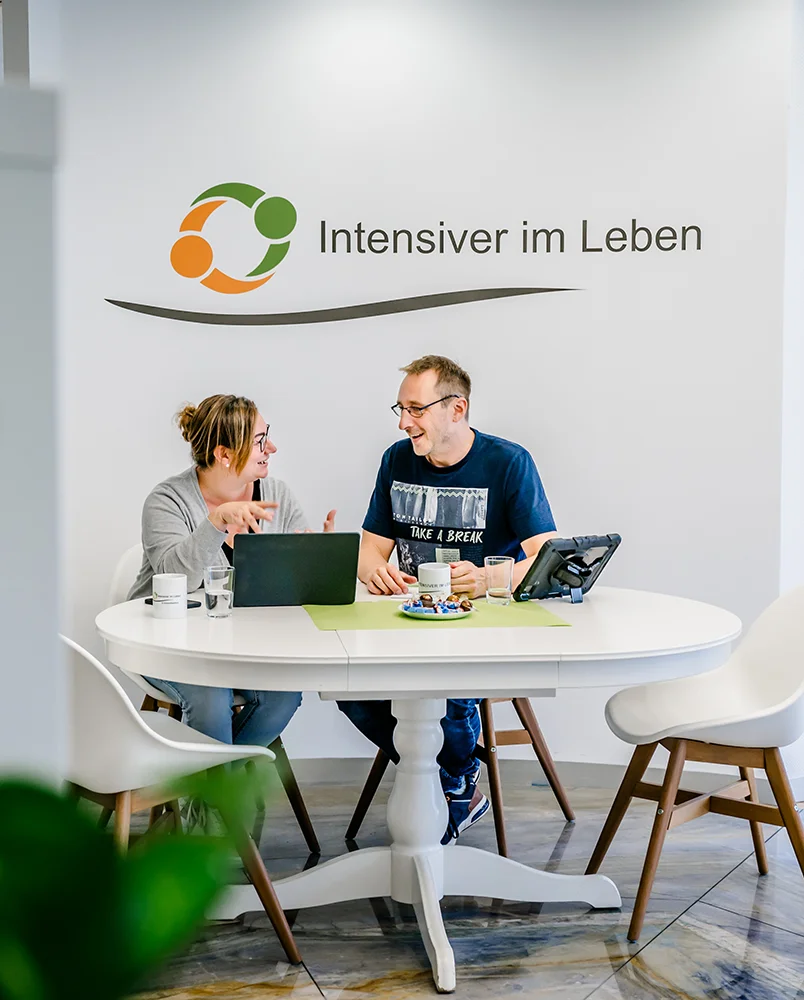 Zwei Mitarbeiter von Intensiver im Leben aus Troisdorf sitzen mit Laptops und Unterlagen an einem runden weißen Tisch, unterhalten sich und lächeln.